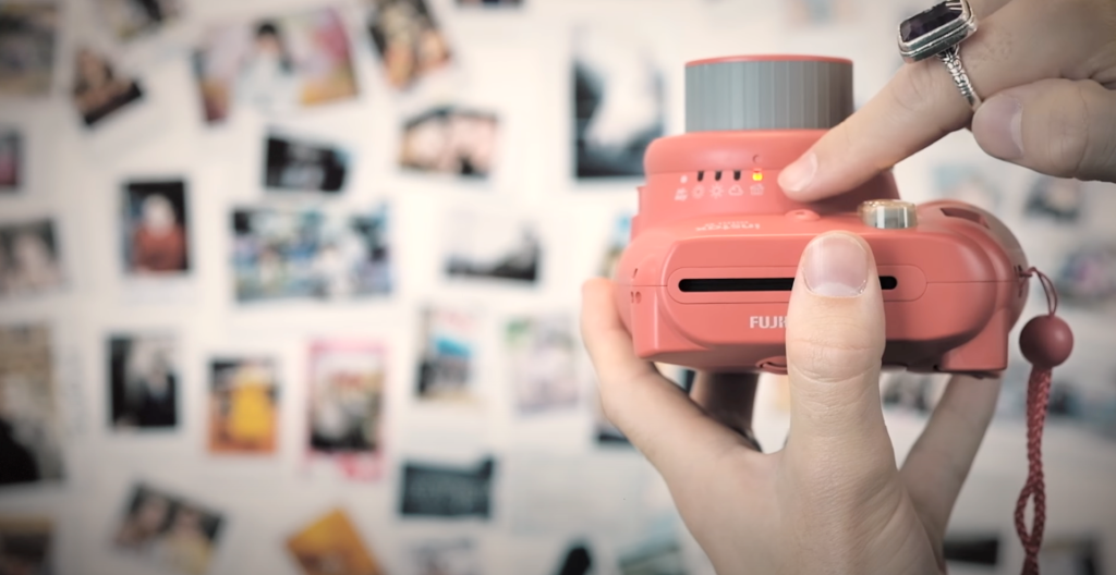 A hand holding a pink Instax camera with the other hand pointing to the adjustment dial, with blurry Polaroid pictures in the background