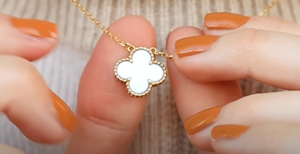 A hand with orange nail polish holding a minimalist necklace with a white pendant.
