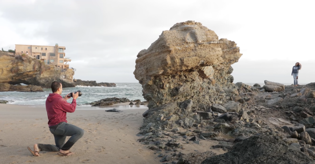 A woman photographing a girl in a red dress at the beach during sunset with large rocks in the background 