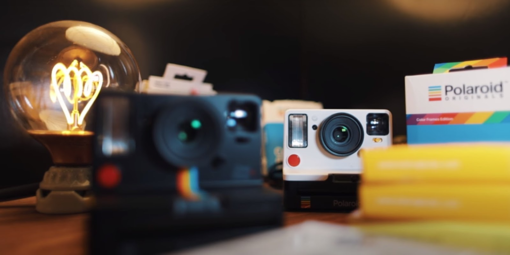 two Polaroid cameras placed on a table and surrounded by a lamp, books, and a package